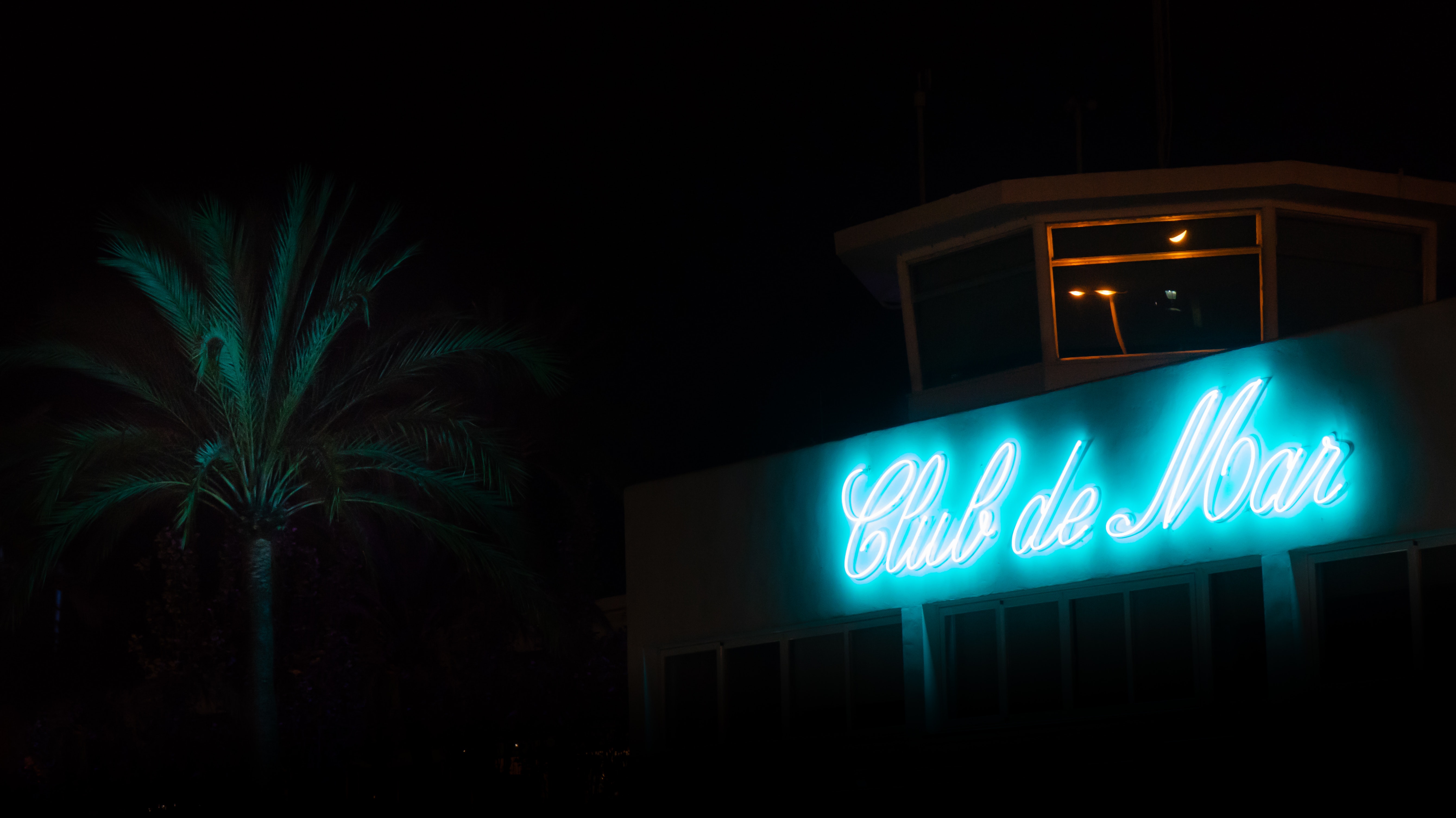 A blue neon sign of Club De Mar near a palm tree
