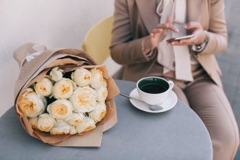 A bouquet of white roses beside a cup of tea in front of a person using a phone