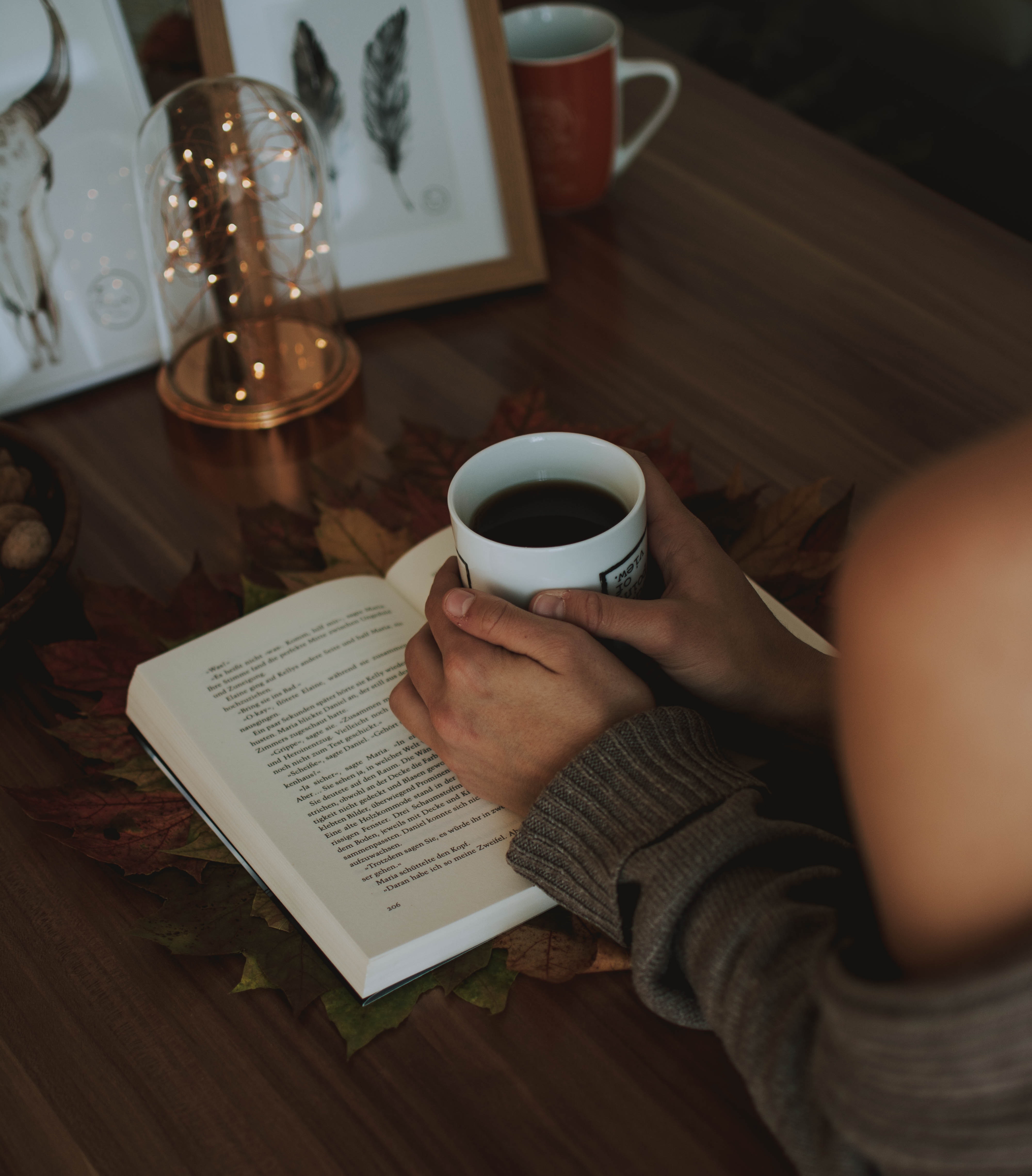 A girl holding a mug of coffee above an opened book on a wooden table