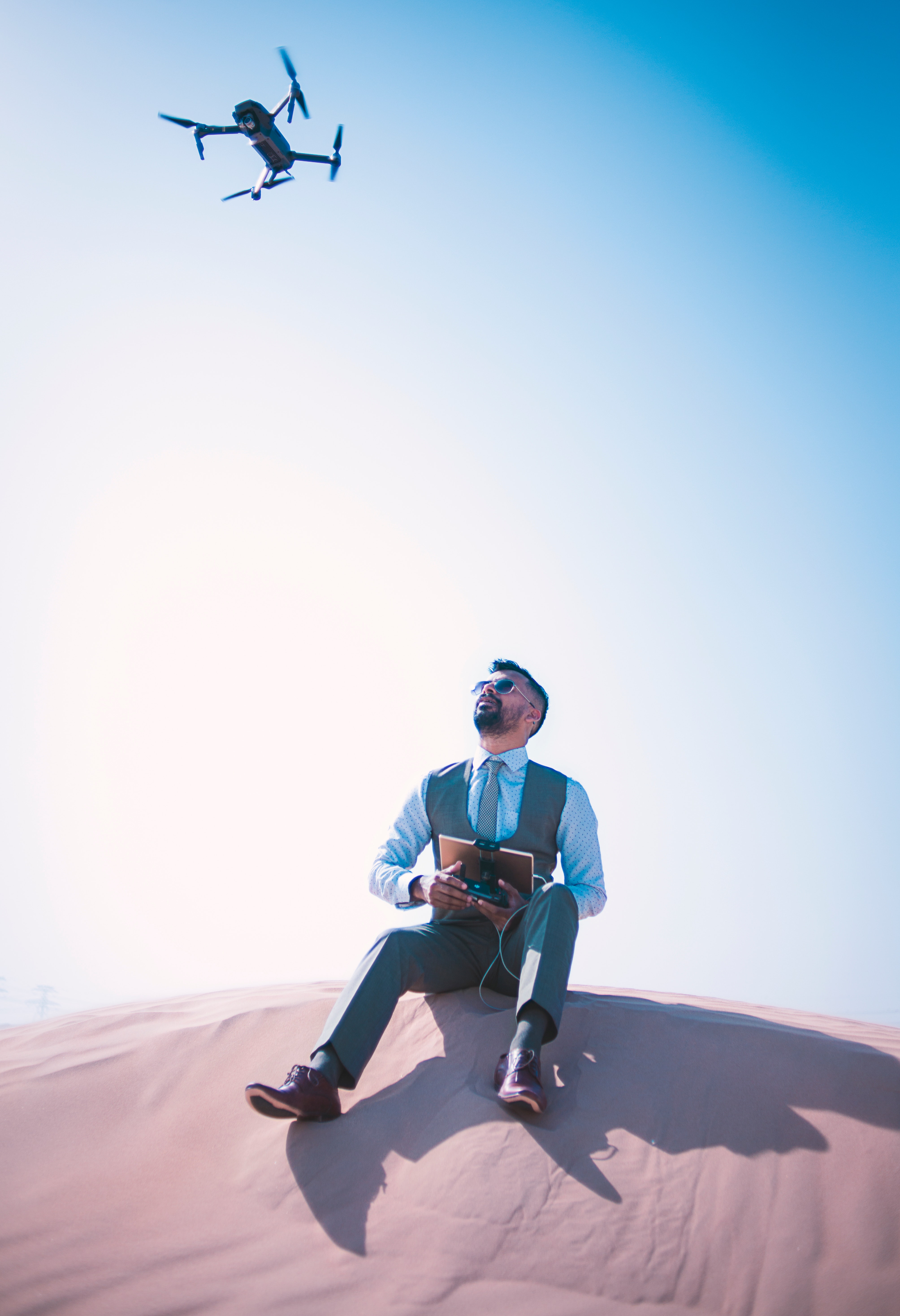 A man sitting on a desert ground looking up and operating a quadcopter