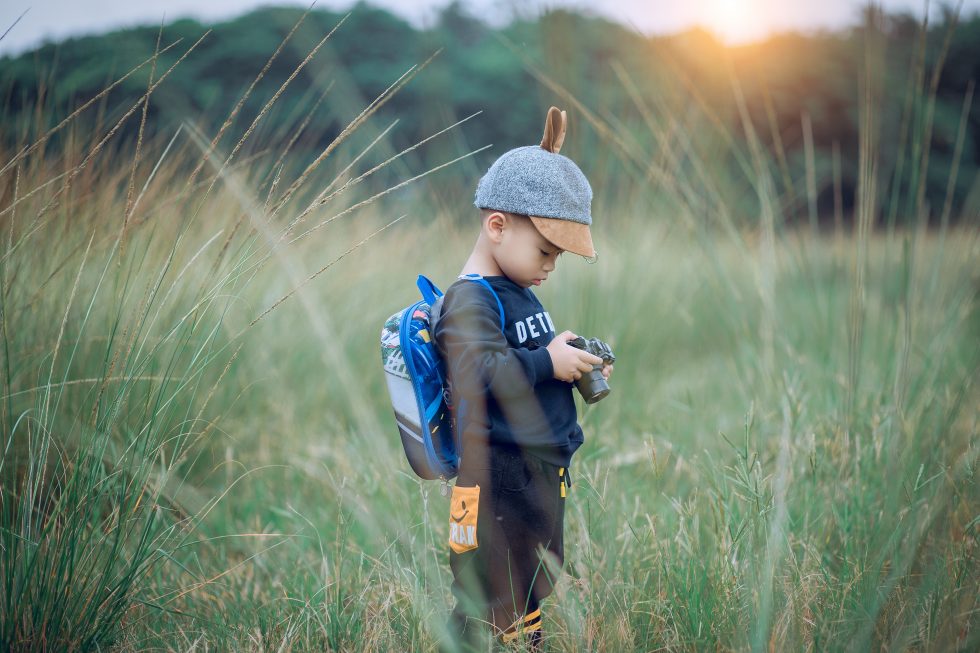 A toddler standing in the middle of the field holding a camera during sunset