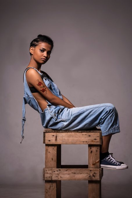 A woman wearing dungarees posing on a wooden stool