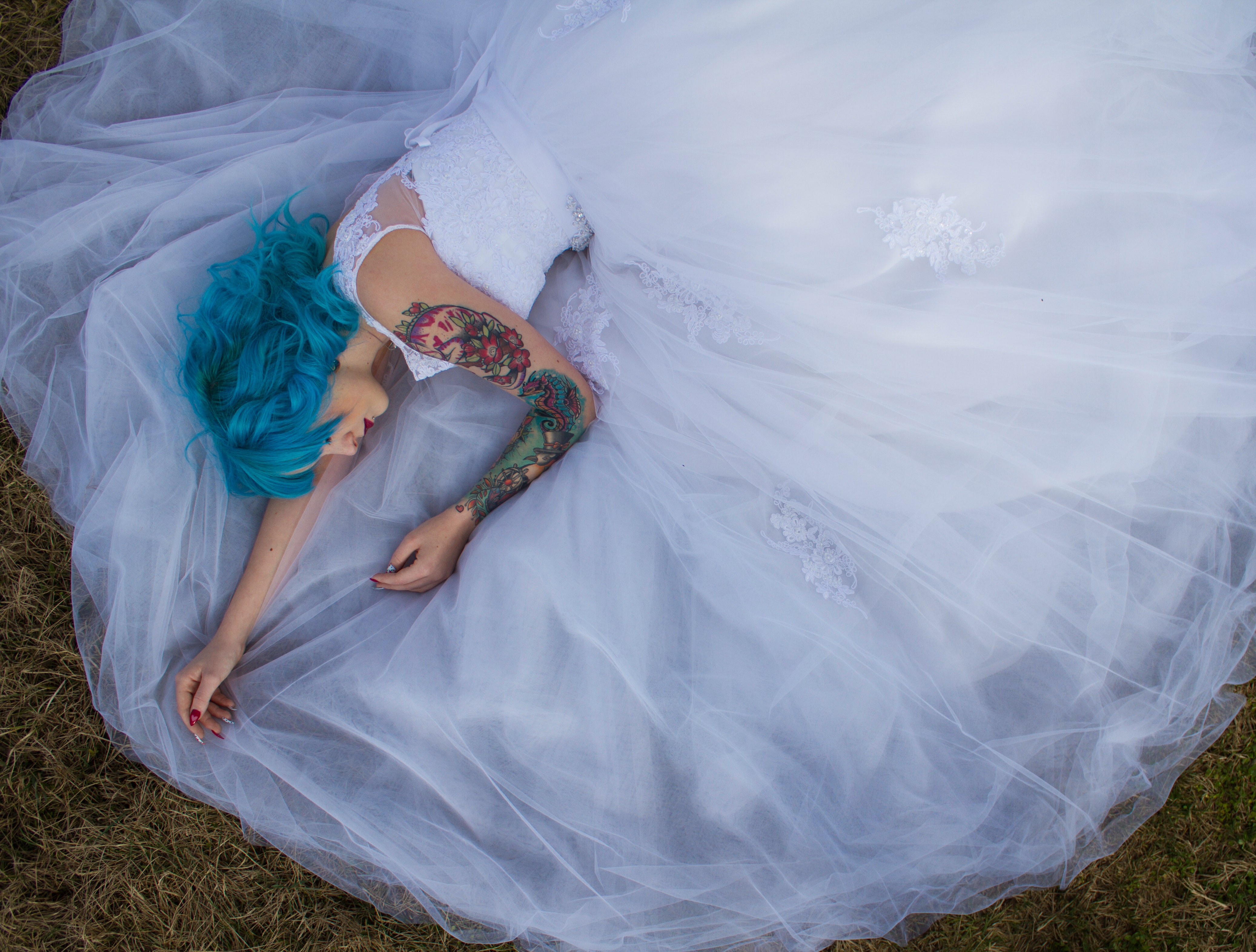A blue-haired woman in a white lace wedding dress lying on green grass