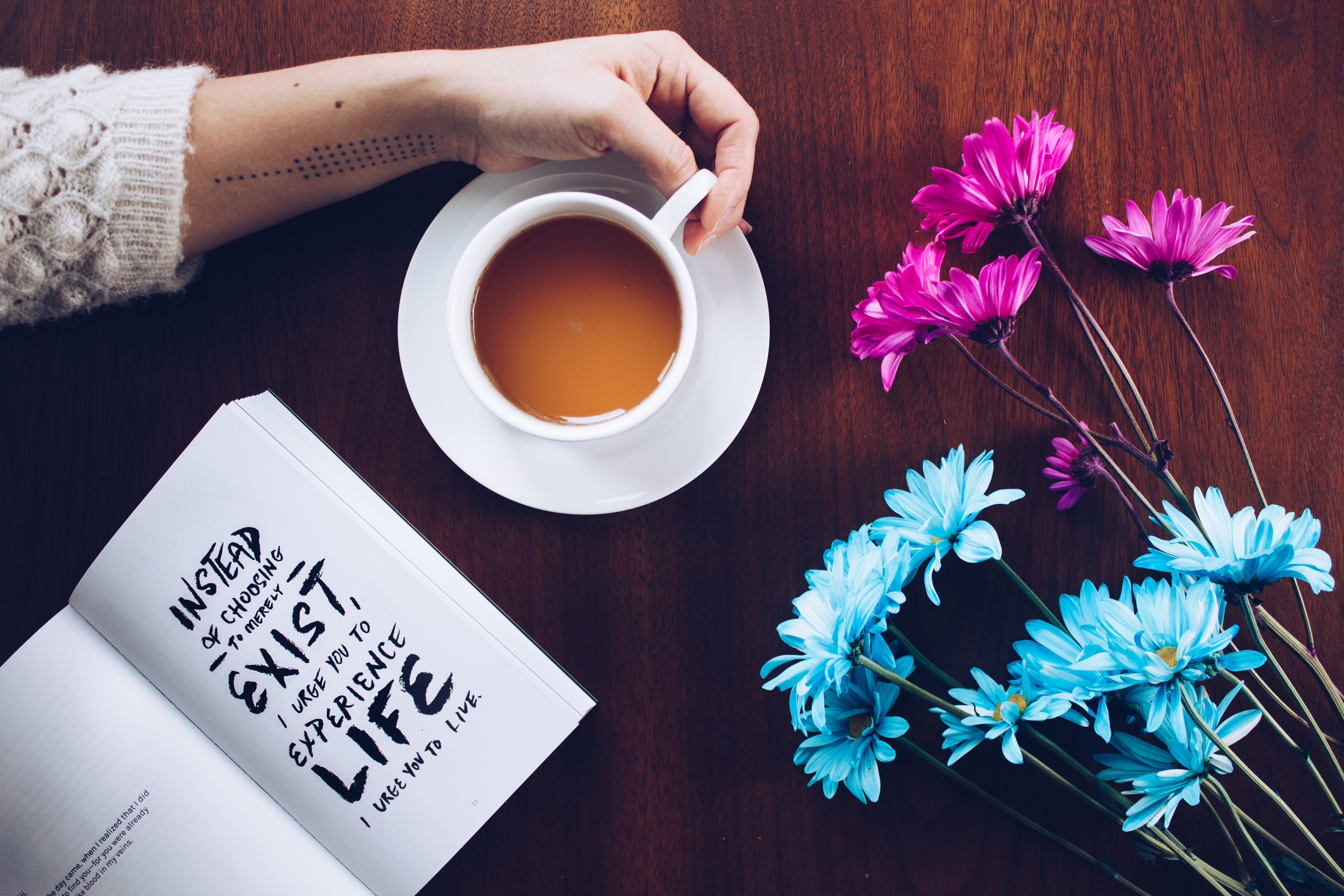 A person holding a cup of coffee beside a book and flowers
