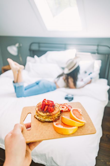A person holding breakfast on a cutting board in front of a woman lying on a bed
