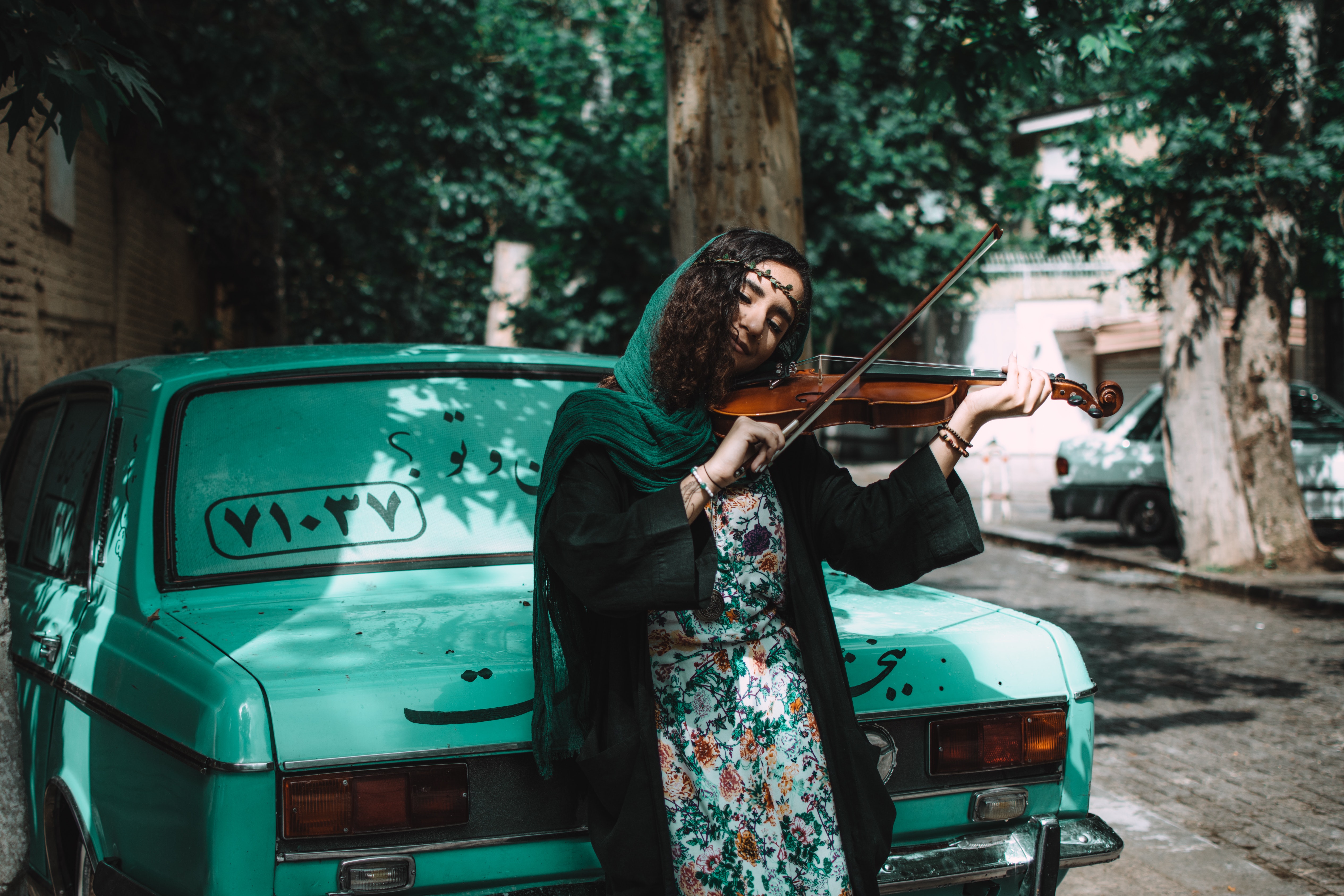 A woman playing the violin while leaning on a green car