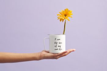 A yellow petaled flower in a white mug in front of a purple wall